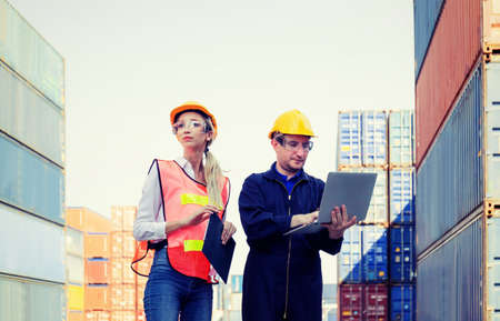 Female foreman safety vest using clipboard checklist and Worker man in hardhat holding laptop for control loading containers box from cargoの写真素材