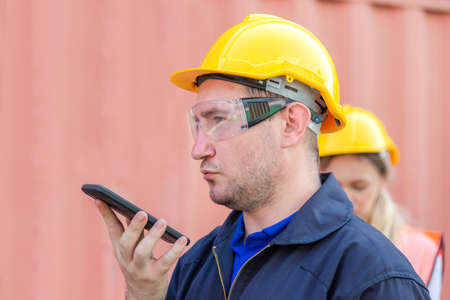 Engineer in the hard hat uses mobile phone, Industrial worker using mobile smartphone in industry containers cargoの写真素材