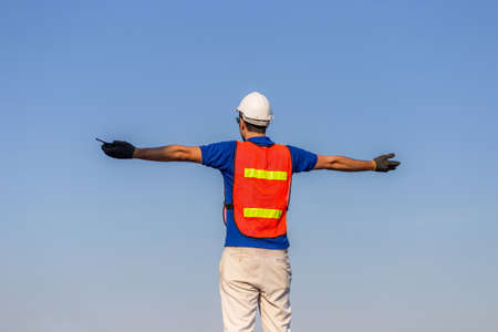 Successful young man with arms wide open, Happy engineer / foreman in hardhat and safety vest talks over blue sky backgroundの写真素材