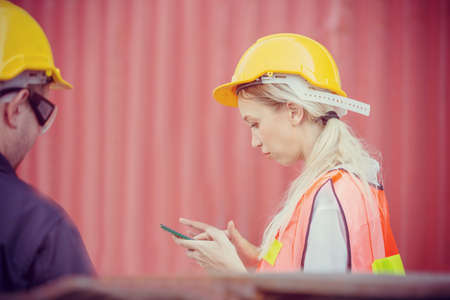Female engineer in the hard hat uses mobile phone, Industrial worker using mobile smartphone in industry containers cargoの写真素材