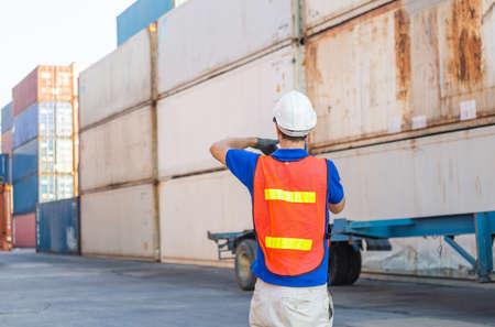 Foreman worker in hard hat and safety vest talks on two-way radio control loading containers box...