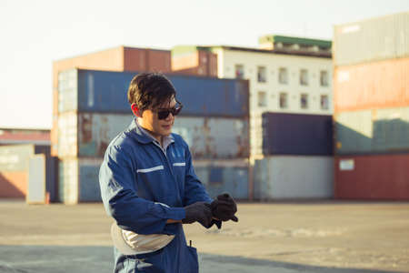 Factory worker man in sunglasses wearing gloves and holding hard hat at cargo containerの写真素材