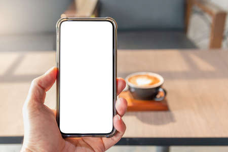 Hand holding smart phone device with empty screen in the coffee shop blurred coffee cup background, Mock up conceptの写真素材