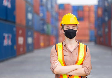 Cheerful female factory worker in hard hat wearing protection face mask against coronavirus with arms crossed as sign of success blurred container box backgroundの写真素材