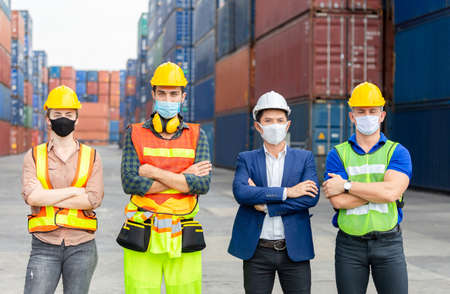 Success Teamwork Concept, Business people engineer and worker team wearing protection face mask against coronavirus with arms crossed as sign of success blurred container box backgroundの写真素材