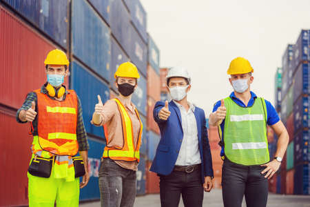 Factory worker and engineer team wearing protection face mask against coronavirus ( COVID-19 ) with giving thumbs up at cargo containersの写真素材