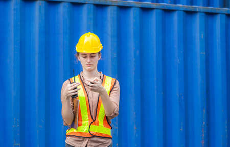 Serious factory worker woman in hard hat standing at containers cargo
