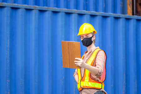 Engineer wearing protection face mask against coronavirus, Woman Foreman in hardhat and safety vest holding clipboard checklist at containers cargoの写真素材
