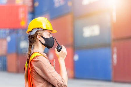 Woman Foreman in hardhat and safety vest holding holding clipboard checklist and talks on two-way radio control loading containers box from cargoの写真素材
