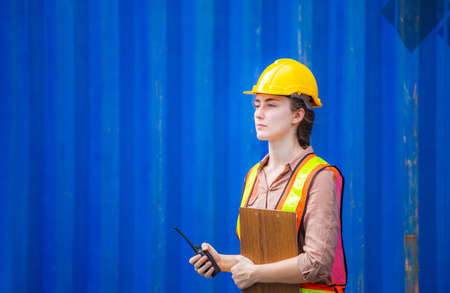 Woman Foreman in hardhat and safety vest holding holding clipboard checklist and two-way radio control loading containers box from cargoの写真素材
