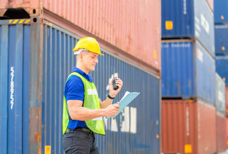 Foreman worker in hardhat and safety vest holding clipboard checklist and talks on two-way radio at...