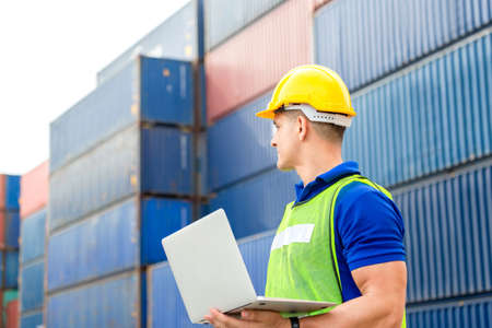 Engineer in hard hat and safety vest holding a laptop, Factory worker man at container cargo