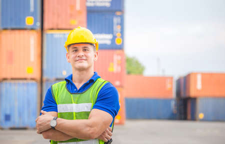 Cheerful factory worker man in hard hat smiling and arms crossed as sign of success with blurred...