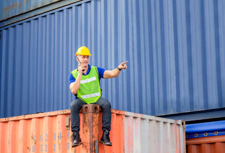 Foreman in hardhat and safety vest sitting on container box talks on two-way radio control loading...