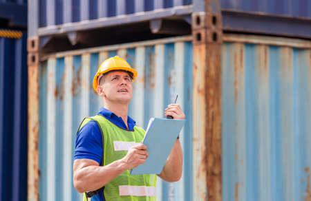 Engineer man in hard hat and safety vest holding clipboard checklist and talks on two-way radio at containers cargoの写真素材