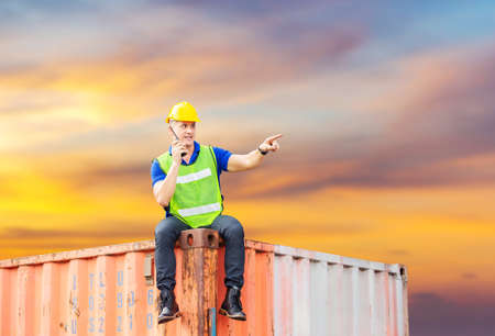 Foreman in hardhat and safety vest sitting on container box with clipping path talks on two-way radio control loading containers box from cargoの写真素材