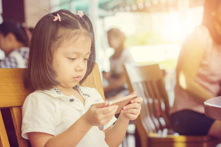 Close up of beautiful little cute girl waiting for her mother to talk with friends in a coffee shop and looking at the smartphoneの写真素材