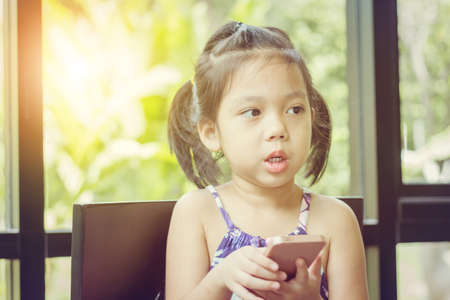 Close up of beautiful little cute girl looking up thinking and holding smartphone, Happy kid concept.の写真素材