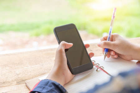 Close up of casual women hand holds mobile smartphone and writeing notes in notebook in cafe, selective focusand blurry background.の写真素材