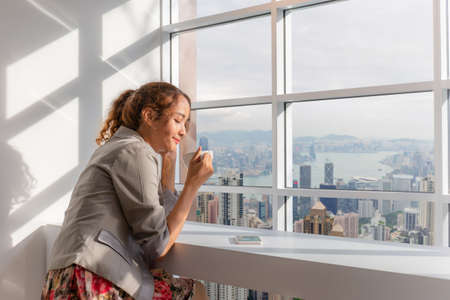 Relaxed young business woman drinking a coffee and watching the city view, enjoying conceptの写真素材