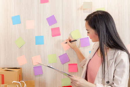 Happy young business woman holding tablet and writing ideas on stick notes on the wall in office
