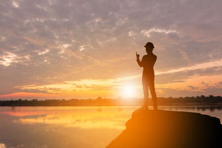 Silhouette of Business Engineer Man Celebration Success Happiness on a Stone Evening Sky Sunset at Lake Background, I am number one and active life Concept.の写真素材