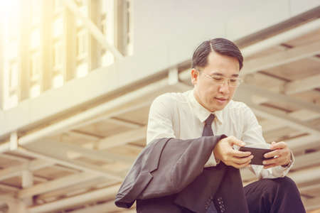 Close up of young business man sitting and checking work on mobile smartphone outside office blurry background.の写真素材