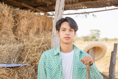 Young farmer man with pitchfork looking camera with blurred bales of pressed straw in rural countryside storage, Smart farmer conceptの写真素材