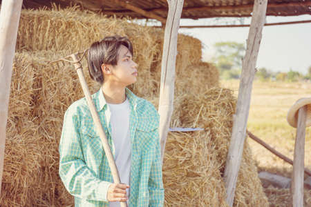 Young farmer man with pitchfork looking the sky with blurred bales of pressed straw in rural countryside storage, Smart farmer conceptの写真素材