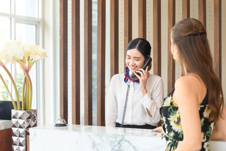 Young woman receptionist talking on the phone at workplace with female travellers standing at reception desk in hotelの写真素材