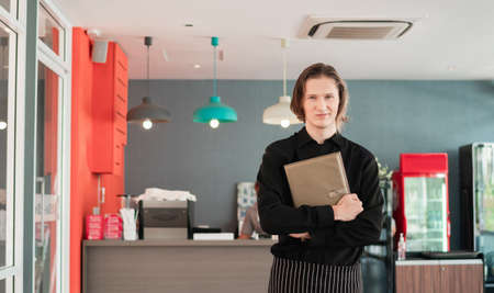Coffee shop worker standing with holding menus serving in bar restaurant. Young man waitress carrying a menu at the counterの写真素材