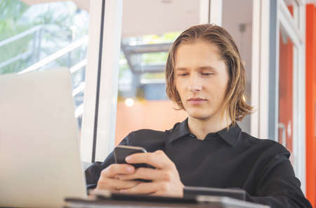 Young man using smartphone and laptop on wooden table in coffee shopの写真素材