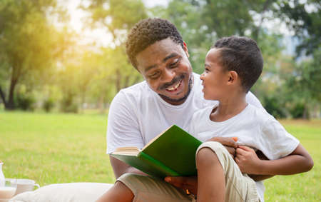Cheerful african american boy and dad having a picnic in the park, Happy son and father reading a book, Happiness family conceptsの写真素材