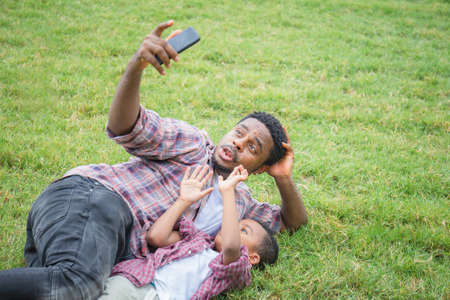 Happy father with little son taking selfie picture with smartphone, Cheerful african american father and sons playing in parkの写真素材