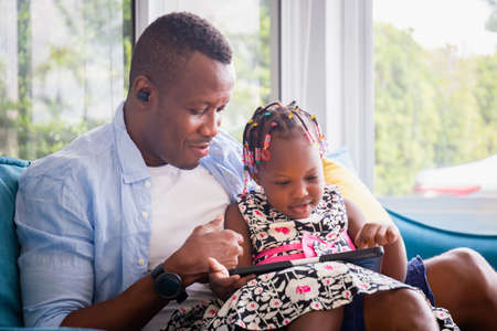 Cheerful african american father and daughter playing in living room, Cute little girl and dad sitting on the sofa and playing on tablet, Happiness family conceptsの写真素材