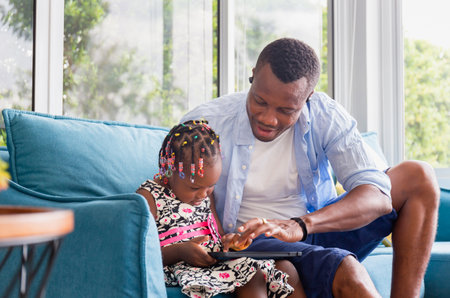 Cheerful african american father and daughter playing in living room, Cute little girl and dad sitting on the sofa and playing on tablet, Happiness family conceptsの写真素材