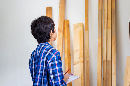 Young carpenter checking woodwork stock at factory storage. Man counting timber beams wood inventory...