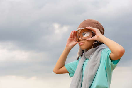 Asian child girl in the old style pilot uniform stands with holding goggles and looking forward, Happy kid playing outdoorsの写真素材