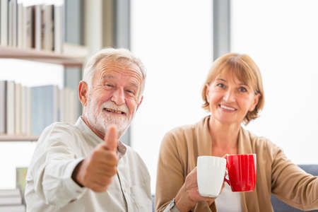 Senior couple inside home during a coffee break, Smiling elderly couple showing thumbs up with holding cups of coffeeの写真素材
