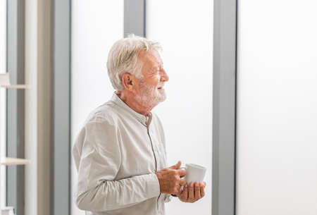 Senior man inside new home during a coffee break, Elderly man standing near window with cups of coffeeの写真素材