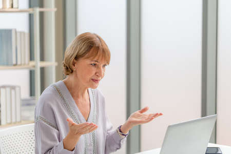 Portrait of woman using laptop and smartphone at home, Mature woman in living room with laptop talking video call on modern computer gadgetの写真素材