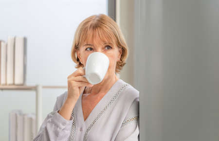Senior woman inside new home during a coffee break, Woman standing near window with cups of coffeeの写真素材