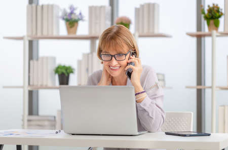 Portrait of woman using laptop and smartphone at home, Mature woman in living room with laptop browsing internet on modern computer gadgetの写真素材