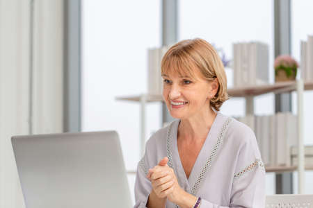 Portrait of woman using laptop and smartphone at home, Mature woman in living room with laptop talking video call on modern computer gadgetの写真素材