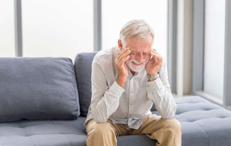 Senior man having headache and touching his head while suffering from a migraine in the living room, Mature man presses a hand to head suffers from unbearable painの写真素材