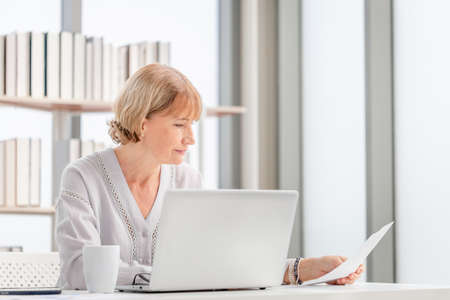 Portrait of woman checking their bills, Mature casual woman reading documents and working on a laptop in living roomの写真素材