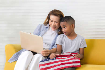 Grandmother and grandchildren playing cheerfully in living room, Woman looking at laptop screen and boy holding usa flagの写真素材