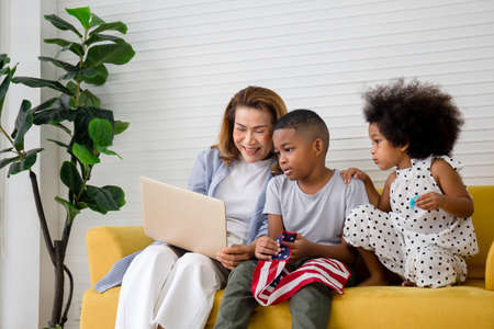 Children and grandmother playing cheerfully in living room, Mother and little children sitting on couch using laptop, boy holding usa flagの写真素材