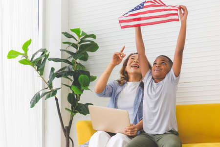 Cheerful mature woman with laptop looking kid boy holding USA flag, Grandmother and grandchildren playing cheerfully in living roomの写真素材