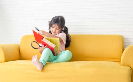 Little kid girl reading book with magnifier on sofa, Happy children playing with magnifying glass in the living roomの写真素材